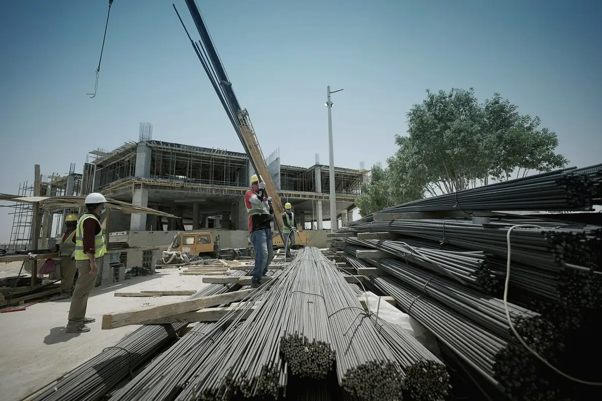 Bundles of reinforcing steel on a construction site with workers and a concrete frame beyond