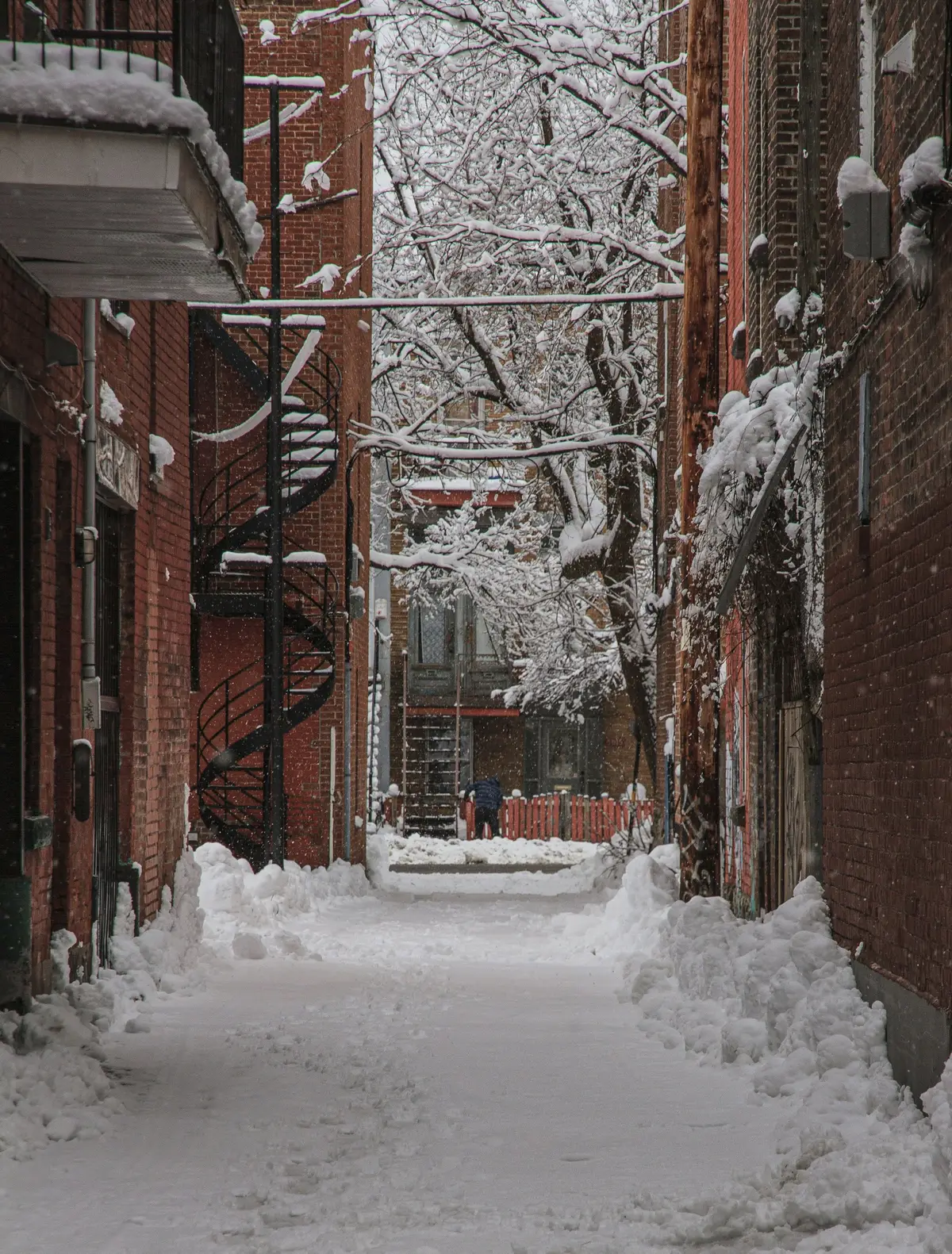 A snow-covered laneway between brick buildings with an exterior spiral stair